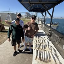 Three men stand by a table with numerous fish by the waterfront.