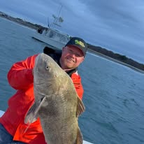 Man in orange jacket holding a large fish on a boat with water in background.